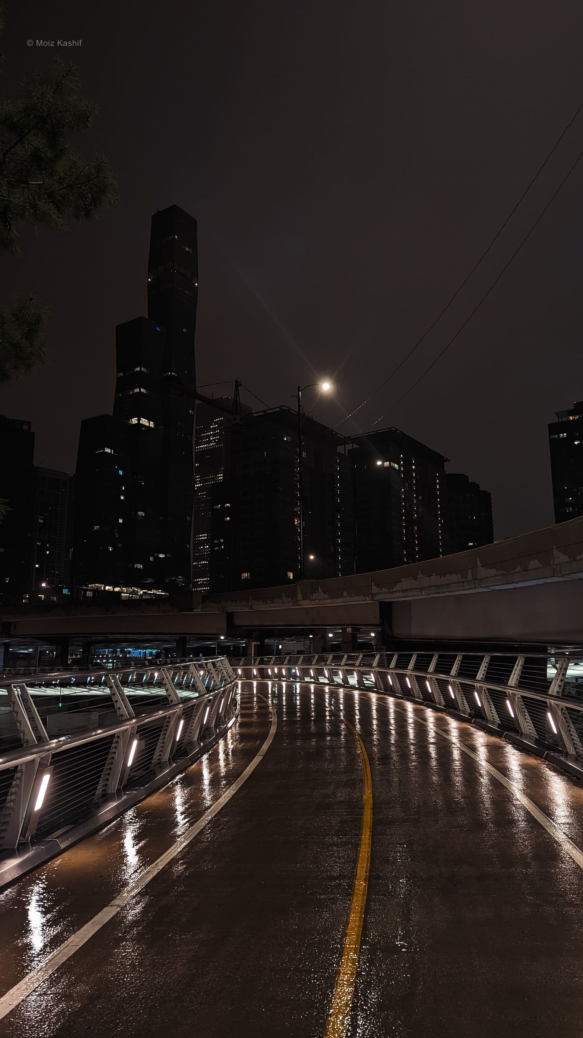 An empty curved walkway reflecting city lights after rain.