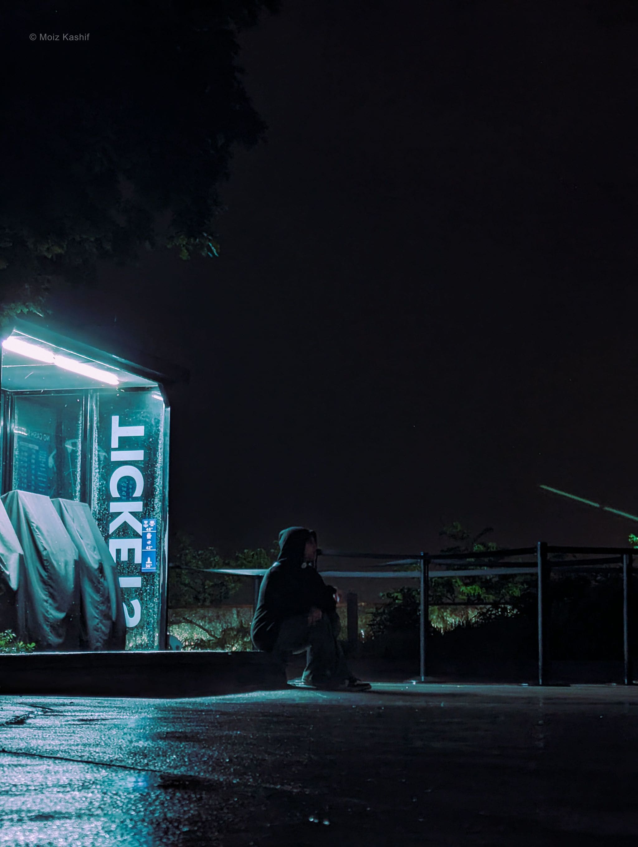 A seated figure beside a glowing ticket booth on a misty lakefront night.