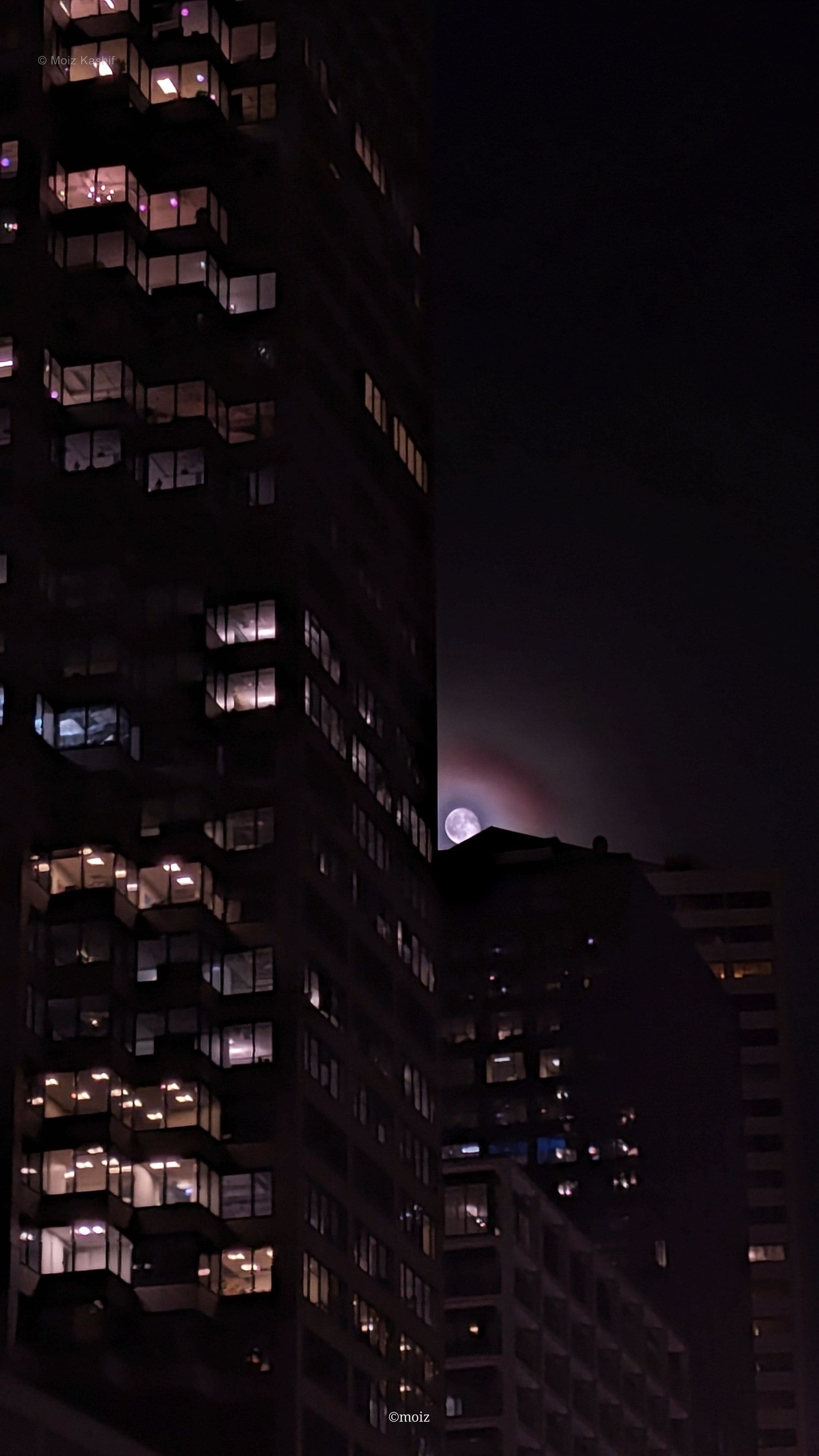 A crescent moon appearing between shadowed apartment towers.