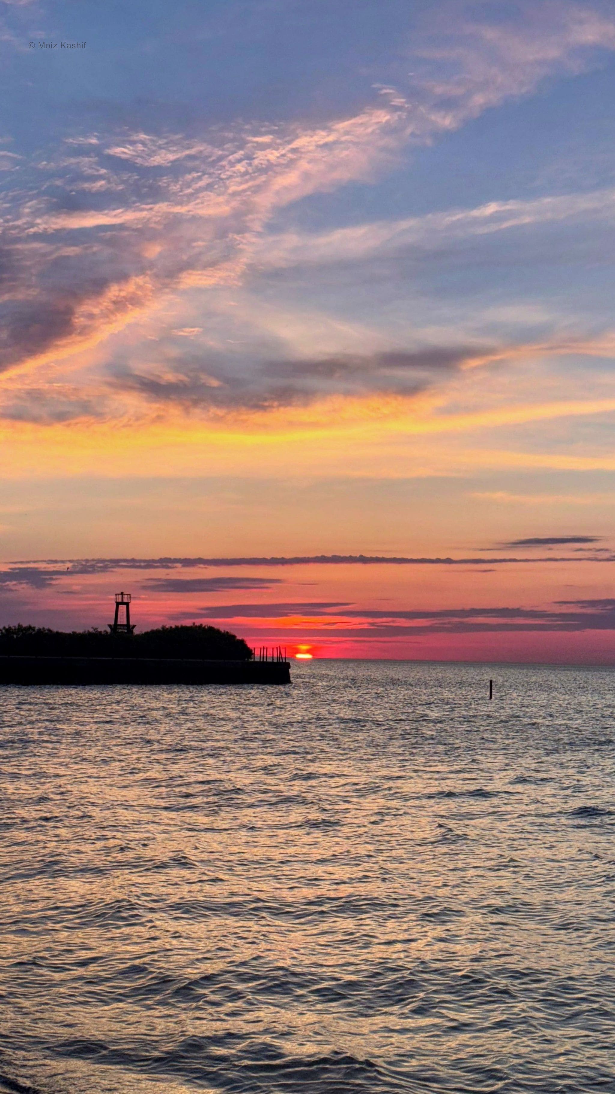 A red sun setting across rippled water beneath streaked clouds.