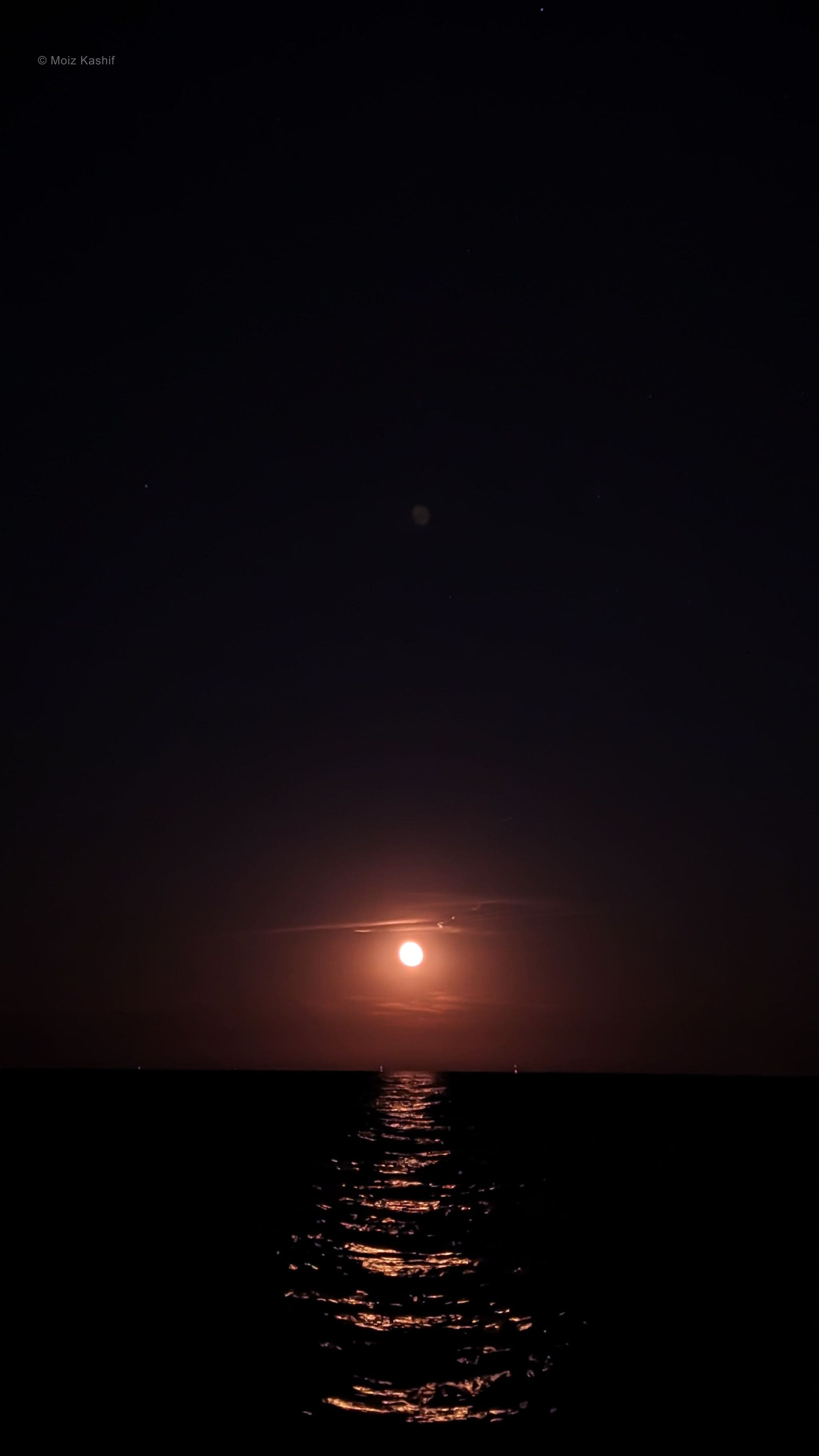 A red crescent moon and its reflection across black lake water.