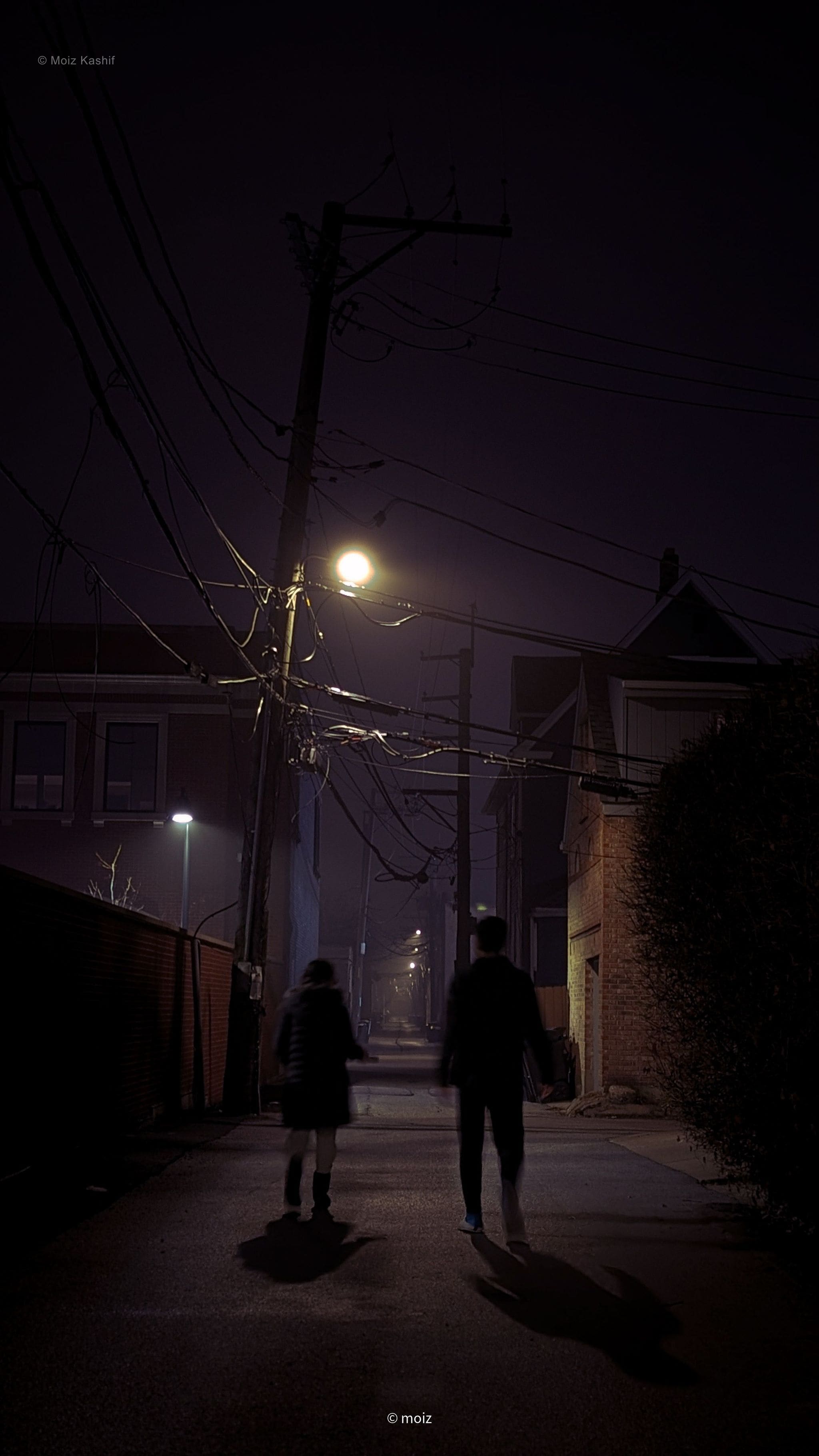 Two people casting long shadows in a narrow night alley.