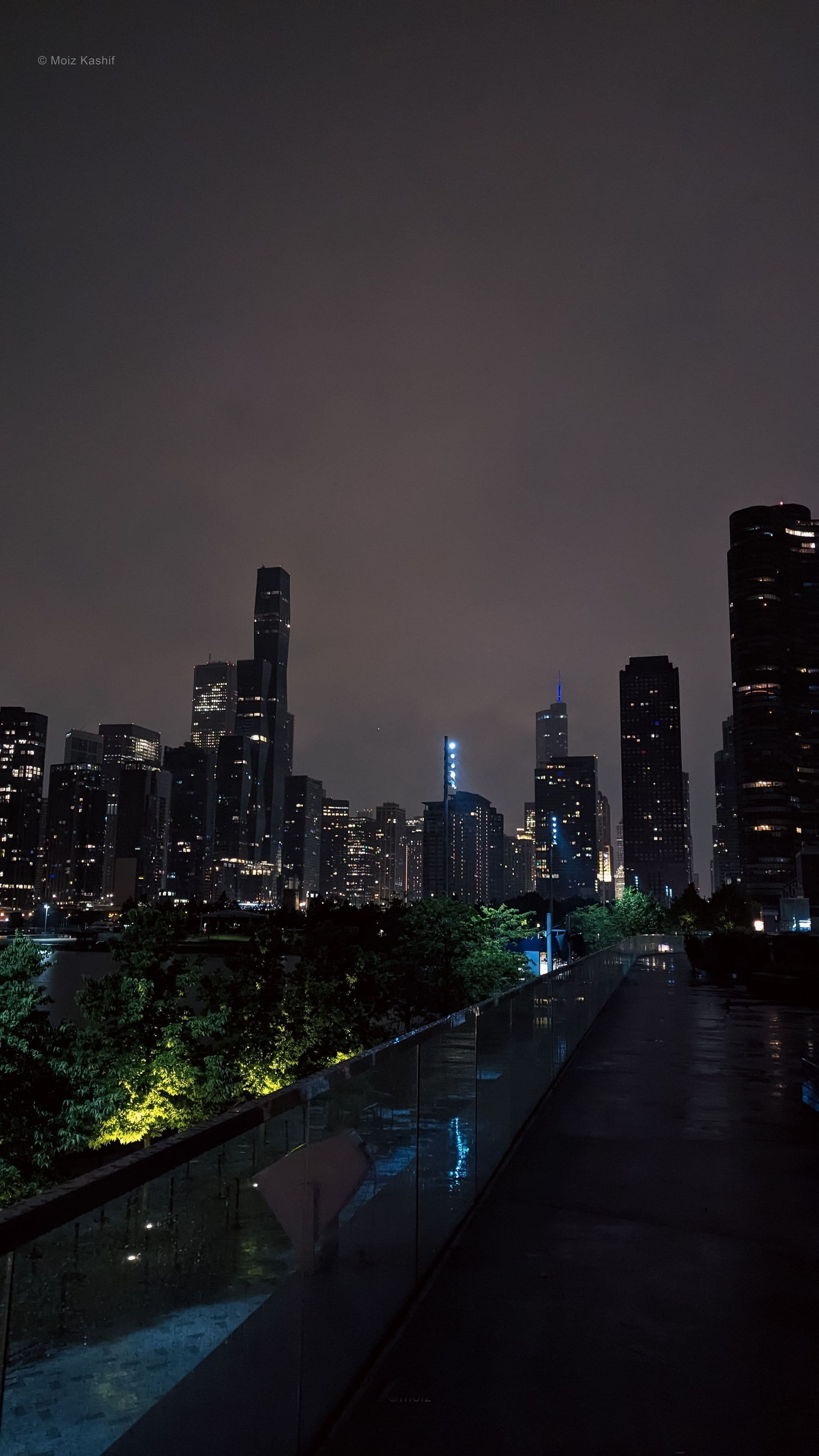 Chicago towers beyond a reflective empty path after rain.