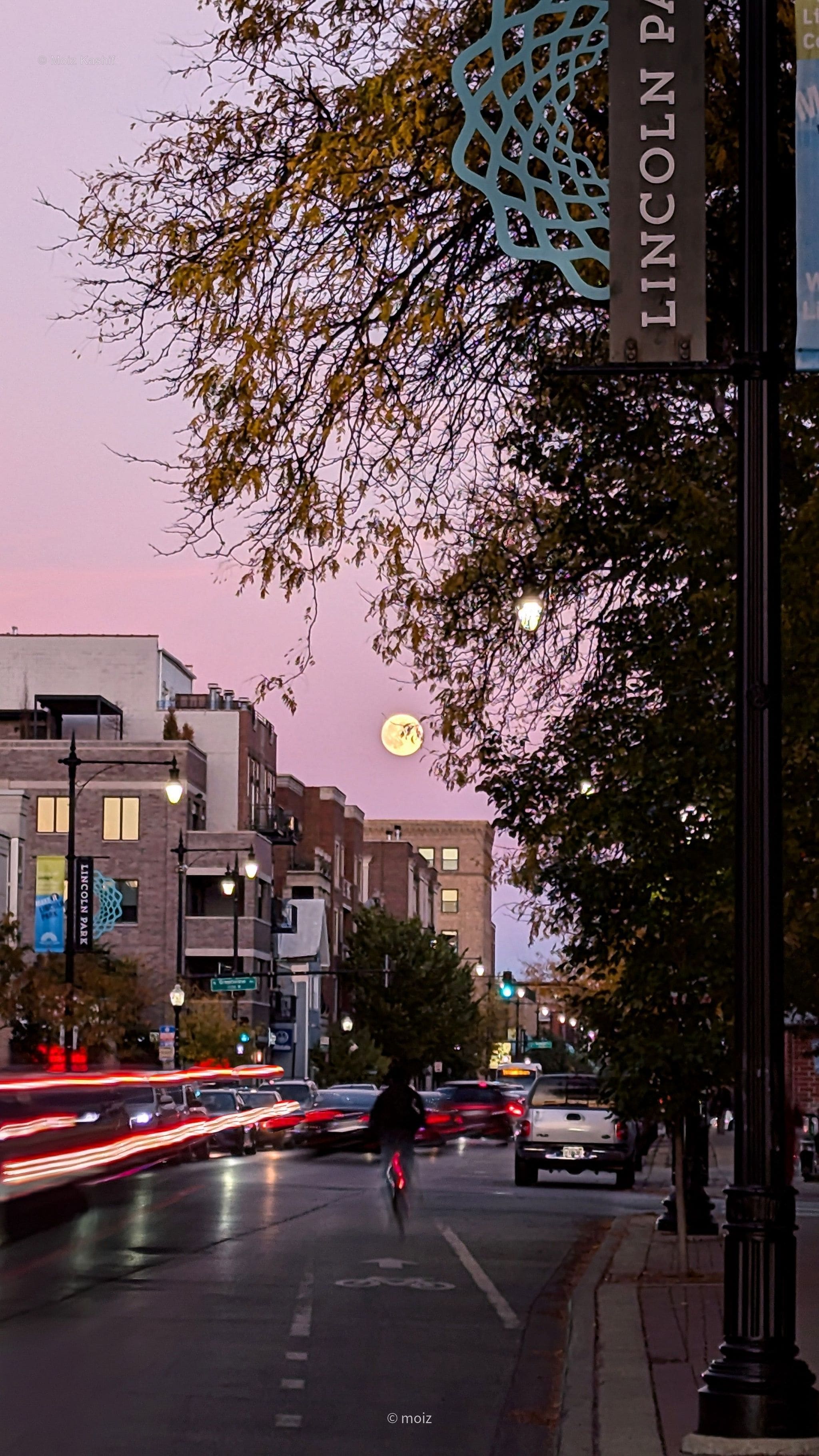 A full moon over Lincoln Avenue with red traffic trails at dusk.