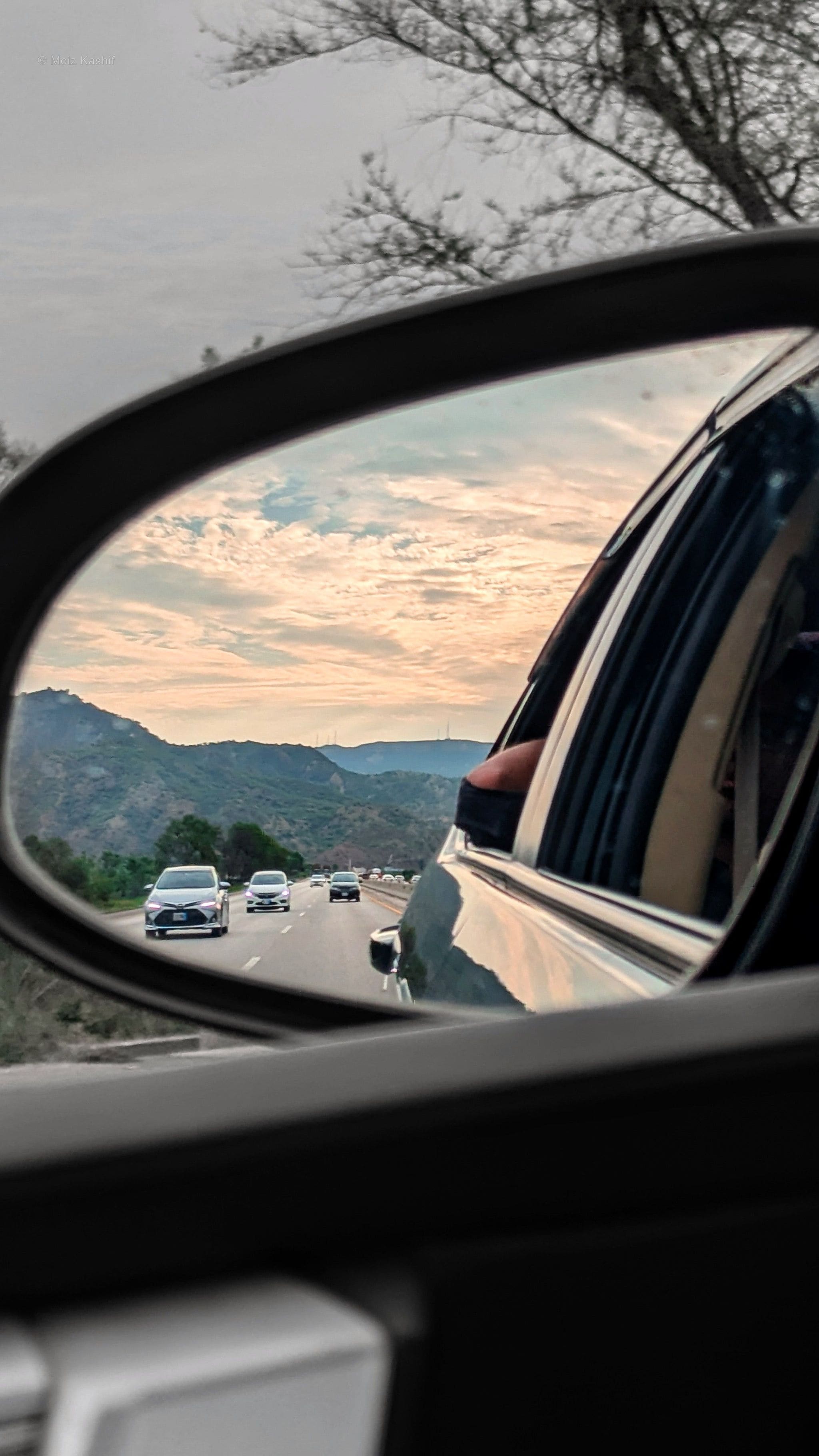 A mountain road and passing cars framed inside a side mirror.