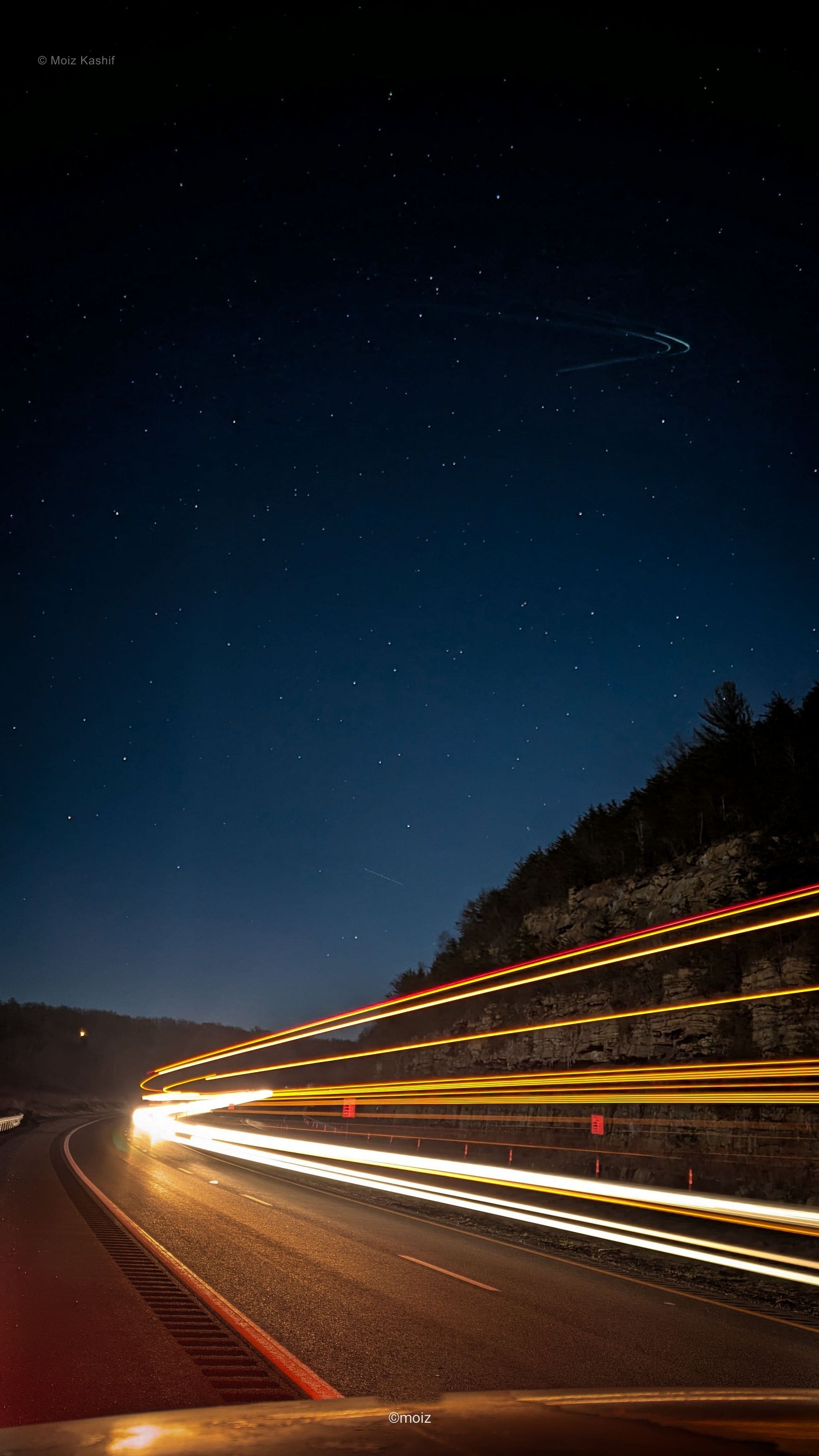 Long white and amber light trails cutting through a mountain road at night.